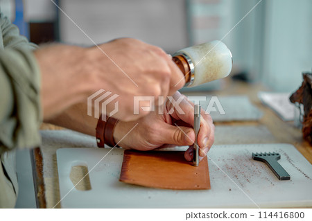 young man's hands punching holes in the leather with a punch. 114416800