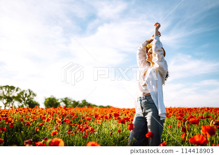 Young girl in hat posing in the poppy field. Portrait of curly girl walking in poppy field at sunset. 114418903