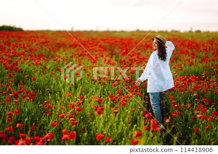 Young girl in hat posing in the poppy field. Portrait of curly girl walking in poppy field at sunset. 114418906