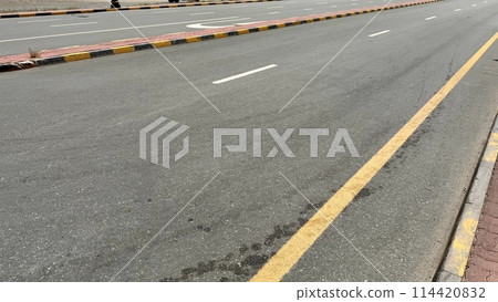 An empty curving auto road with distinct yellow restriction lines, white lane markings and a sidewalk edge 114420832