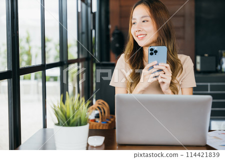 Happy lifestyle female smiling using smartphone in coffee shop, young business woman working with laptop computer she holding smart mobile phone looking out of windows at cefa for texting messages 114421839
