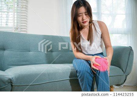 Asian young woman sitting on sofa and feeling suffering knee pain in living room at home, female massage her knee leg pain with painful face, Health bone problems self physiotherapy 114423255