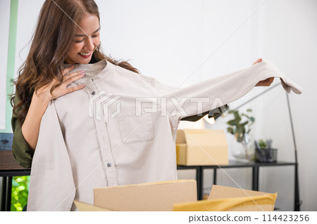 Happy Asian woman customer opening box and shirt against body new clothes, Excited smiling female shopper unboxing cardboard box package, shopping online from home, delivery shipping concept 114423256