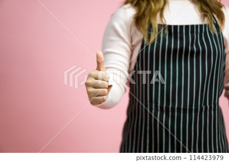 Young woman in pink sweater and striped apron give a thumbs up gesture. Portrait on pink background with studio light. Young woman in pink sweater and striped apron give a thumbs up gesture. Portrait on pink background with studio light. 114423979