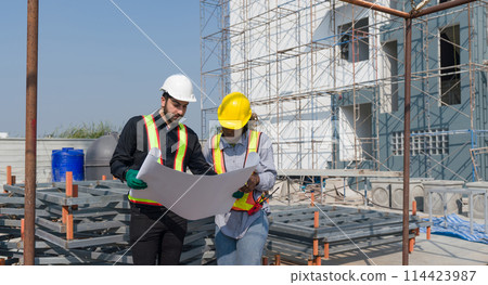 Two construction worker looking at a floor plan, standing in front of a large building site with helmet and safety gear. Day time work safety check. Work environment at the site of housing projects. Two construction worker looking at a floor plan, standing in front of a large building site with helmet and safety gear. Day time work safety check. Work environment at the site of housing projects. 114423987