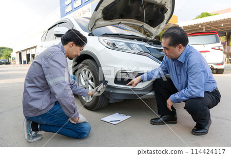 Young mechanic in uniform with protective gloves, taking photo of a vehicle with a smartphone. Some paperwork is on the ground, suggesting that they are following a checklist or taking notes. 114424117