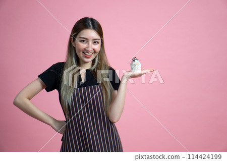 Young asian woman with an apron in celebratory or festive mood holding cupcake, decorated with white frosting and chocolate chip. Portrait on pink background with studio light. 114424199