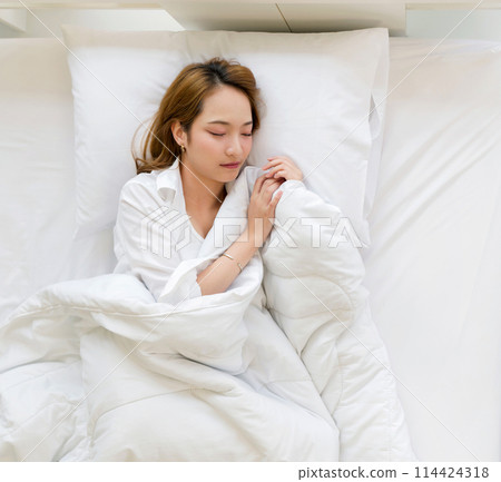 Young asian woman in white pajama, sleep and sweet dream lying on bed with soft pillows and a comfortable duvet wrapped around. Top view 114424318