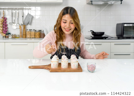 Young woman in an apron holding spoon with multicolor frosting, decorating cupcake on a wooden board, adding a colorful touch to the cupcakes' presentation. Young woman in an apron holding spoon with multicolor frosting, decorating cupcake on a wooden board, adding a colorful touch to the cupcakes' presentation. 114424322