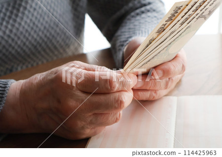 An elderly woman's hand holding banknotes and a bankbook on a desk An elderly woman's hand holding banknotes and a bankbook on a desk 114425963