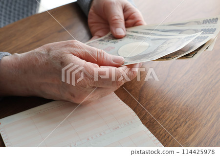 An elderly woman's hand holding banknotes and a bankbook on a desk An elderly woman's hand holding banknotes and a bankbook on a desk 114425978