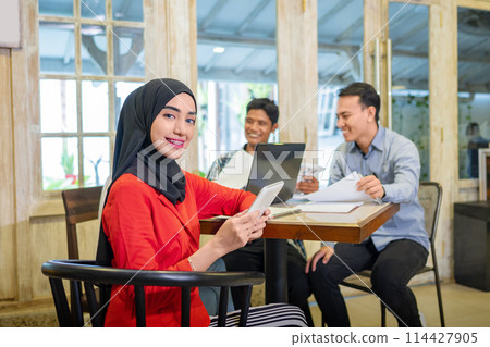 Indonesian young pretty entrepreneur muslim woman smile at the camera while holding smartphone, against business meeting with clients on a cafe. 114427905