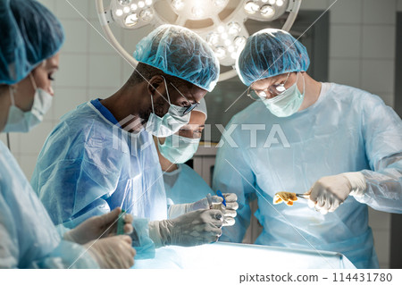 young african assistant helping the doctor to cut the body of patient 114431780