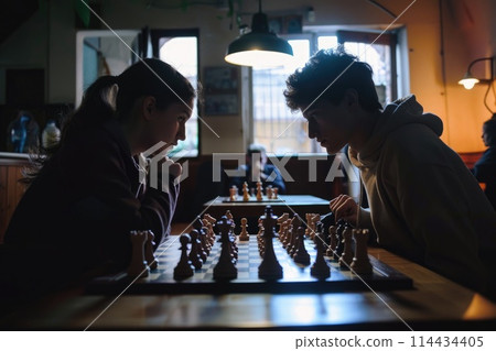 Two young individuals deeply engrossed in a strategic game of chess, competition under soft lighting Two young individuals deeply engrossed in a strategic game of chess, competition under soft lighting 114434405