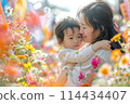 Joyful mother hugging her delighted little girl surrounded by lush garden flowers, celebrating togetherness 114434407