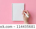 A woman's hand holds a white pen over a blank notebook on a soft pink background 114435681