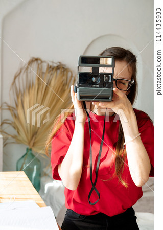A young woman in a red T-shirt holds a polaroid in her hands and takes a photo 114435933