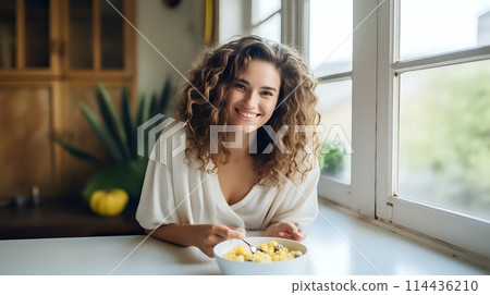 A beautiful young woman is eating a bowl of cereal in the kitche 114436210