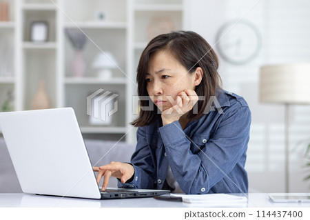 Asian woman looking intensely at a laptop screen while working from home. She epitomizes concentration and dedication in home office, remote work lifestyle and the challenges Asian woman looking intensely at a laptop screen while working from home. She epitomizes concentration and dedication in home office, remote work lifestyle and the challenges 114437400