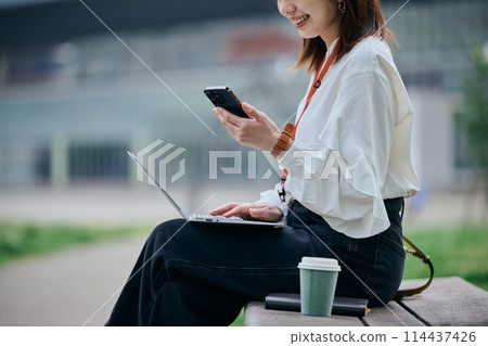 A young woman operating a computer and smartphone in the park 114437426