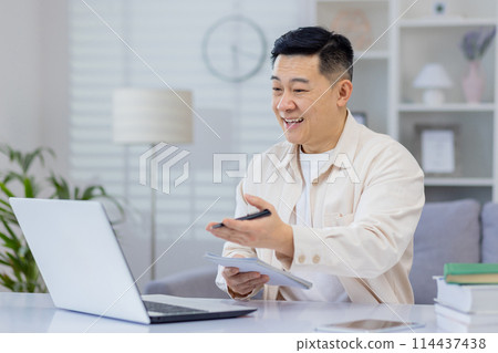Asian man interacts with a webinar on his laptop at home, expressing happiness and engagement. This image captures the joy of lifelong learning and the convenience of online education. 114437438