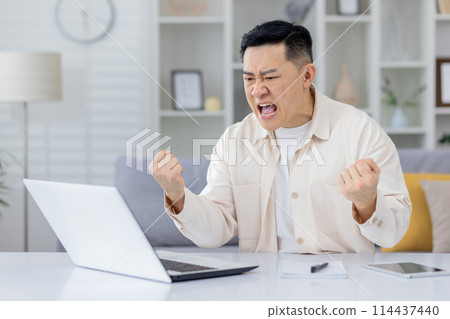 An adult man expressing frustration and anger while working from home, upset with laptop screen. This image captures an intense moment of emotional stress in a remote work environment. 114437440
