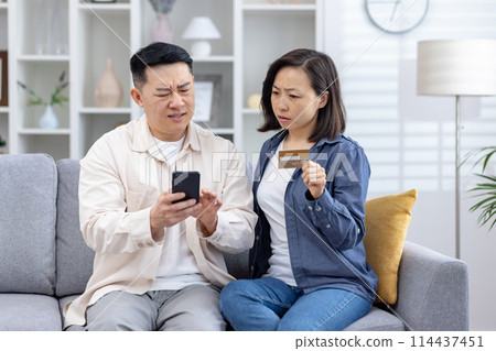 Asian couple looks distressed as they uncover unauthorized charges on their credit card phone, man confusion while the woman, holding the card, seems anxious. stress, frustration of financial fraud. Asian couple looks distressed as they uncover unauthorized charges on their credit card phone, man confusion while the woman, holding the card, seems anxious. stress, frustration of financial fraud. 114437451
