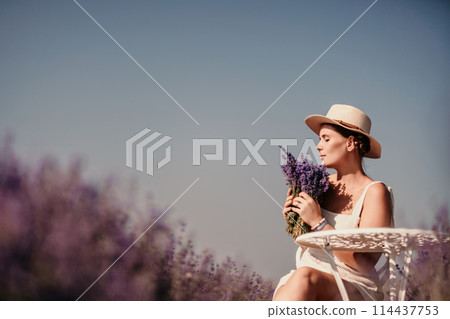 woman sitting in a field of lavender and wearing a straw hat. She is smiling and holding a bouquet of flowers. Scene is peaceful and serene, as the woman is surrounded by the beauty of nature 114437753