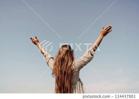 A woman with long hair is standing on the beach, looking up at the sky. She is wearing a dress and she is happy. 114437801