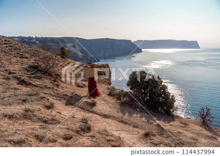 A woman in a red dress stands on a rocky cliff overlooking the ocean. She is wearing a straw hat and she is enjoying the view. 114437994