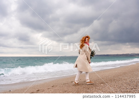 Redhead woman Christmas tree sea. Christmas portrait of a happy redhead woman walking along the beach and holding a Christmas tree in her hands. Dressed in a light coat, white suit and red mittens. 114437995