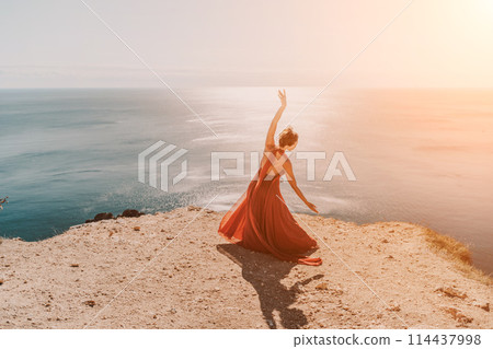 Woman red dress sea. Female dancer in a long red dress posing on a beach with rocks on sunny day. Girl on the nature on blue sky background. Woman red dress sea. Female dancer in a long red dress posing on a beach with rocks on sunny day. Girl on the nature on blue sky background. 114437998