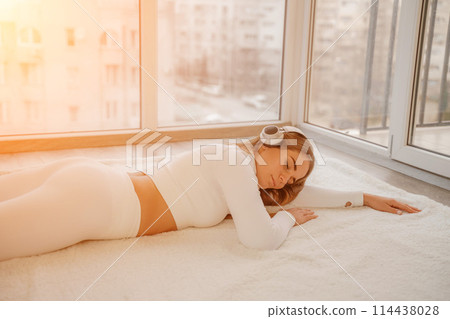Side view portrait of relaxed woman listening to music with headphones lying on carpet at home. She is dressed in a white tracksuit. 114438028