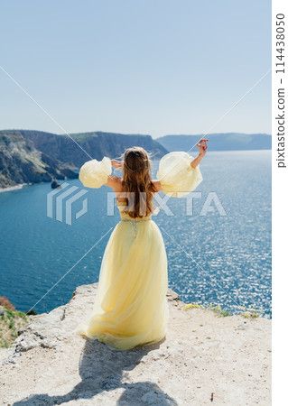 Woman in a yellow dress on the sea. Side view Young beautiful sensual woman in yellow long dress posing on a rock high above the sea at sunset. Girl in nature against the blue sky 114438050