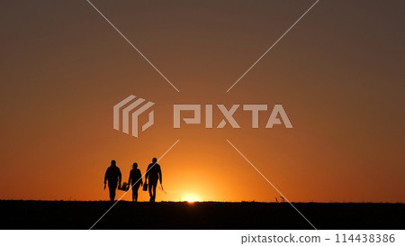 At sunset, three farmers walk through a field, carrying their equipment. 114438386