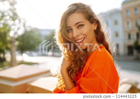 Young woman posing against backdrop of urban landscape in bright orange dress. Concept of lifestyle. 114443225