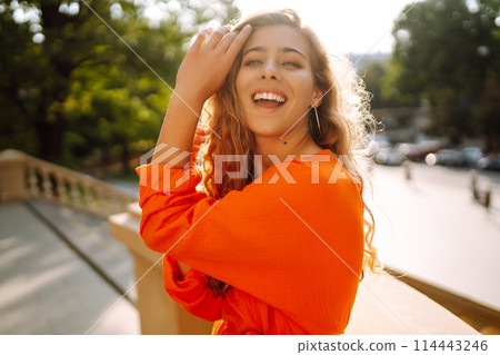Young woman posing against backdrop of urban landscape in bright orange dress. Concept of lifestyle. Young woman posing against backdrop of urban landscape in bright orange dress. Concept of lifestyle. 114443246