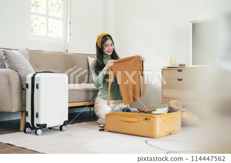 Happy Asian young woman packing suitcase at home. Preparing for summer holiday vacation abroad travel. 114447562