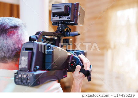 A gray-haired man holds a large professional video camera in his hands. 114456229