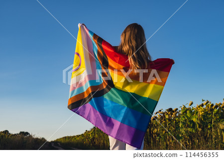 Girl showing her gender identity. Young woman stand with pride flag Rainbow LGBTQIA flag made from silk material on field background. Symbol of LGBTQ pride month. Equal rights. Peace Girl showing her gender identity. Young woman stand with pride flag Rainbow LGBTQIA flag made from silk material on field background. Symbol of LGBTQ pride month. Equal rights. Peace 114456355