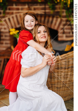 Mother and daughter cuddling in a room by the fireplace in elegant dresses Mother and daughter cuddling in a room by the fireplace in elegant dresses 114457015