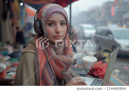 A woman in a hijab and headphones sits at a table with a laptop 114460904