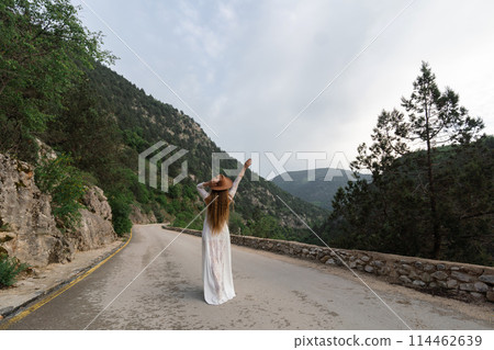 A woman in a white dress is standing on a road, with a hat on her head 114462639