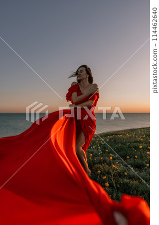 woman red dress standing grassy hillside. The sun is setting in the background, casting a warm glow over the scene. The woman is enjoying the beautiful view and the peaceful atmosphere. 114462640