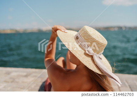 A woman in a swimsuit sits with her back holding a hat, looks at the ocean, sunny day, relaxes. 114462703
