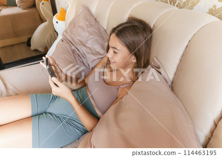 A girl is laying on a bed with a cell phone in her hand. She is smiling and she is enjoying herself. A girl is laying on a bed with a cell phone in her hand. She is smiling and she is enjoying herself. 114463195