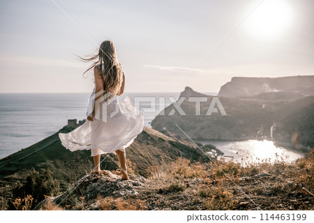 A woman stands on a hill overlooking the ocean. She is wearing a white dress and has long hair. The scene is serene and peaceful, with the sun shining brightly in the background. 114463199