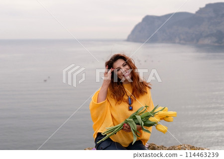 Portrait happy woman woman with long hair against a background of mountains and sea. Holding a bouquet of yellow tulips in her hands, wearing a yellow sweater 114463239