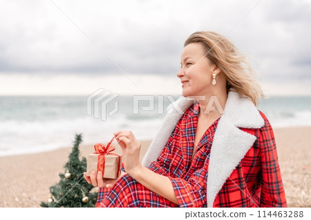Lady in plaid shirt holding a gift in his hands enjoys beach with Christmas tree. Coastal area. Christmas, New Year holidays concep 114463288