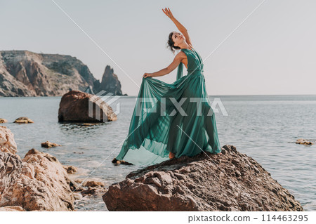 Woman green dress sea. Woman in a long mint dress posing on a beach with rocks on sunny day. Girl on the nature on blue sky background. Woman green dress sea. Woman in a long mint dress posing on a beach with rocks on sunny day. Girl on the nature on blue sky background. 114463295